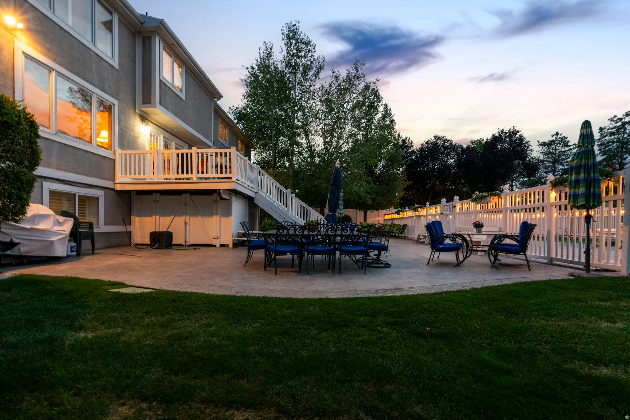 Expansive stamped concrete patio with multiple seating areas, a white picket fence, a spacious upper deck with stairs, and a well-maintained lawn
