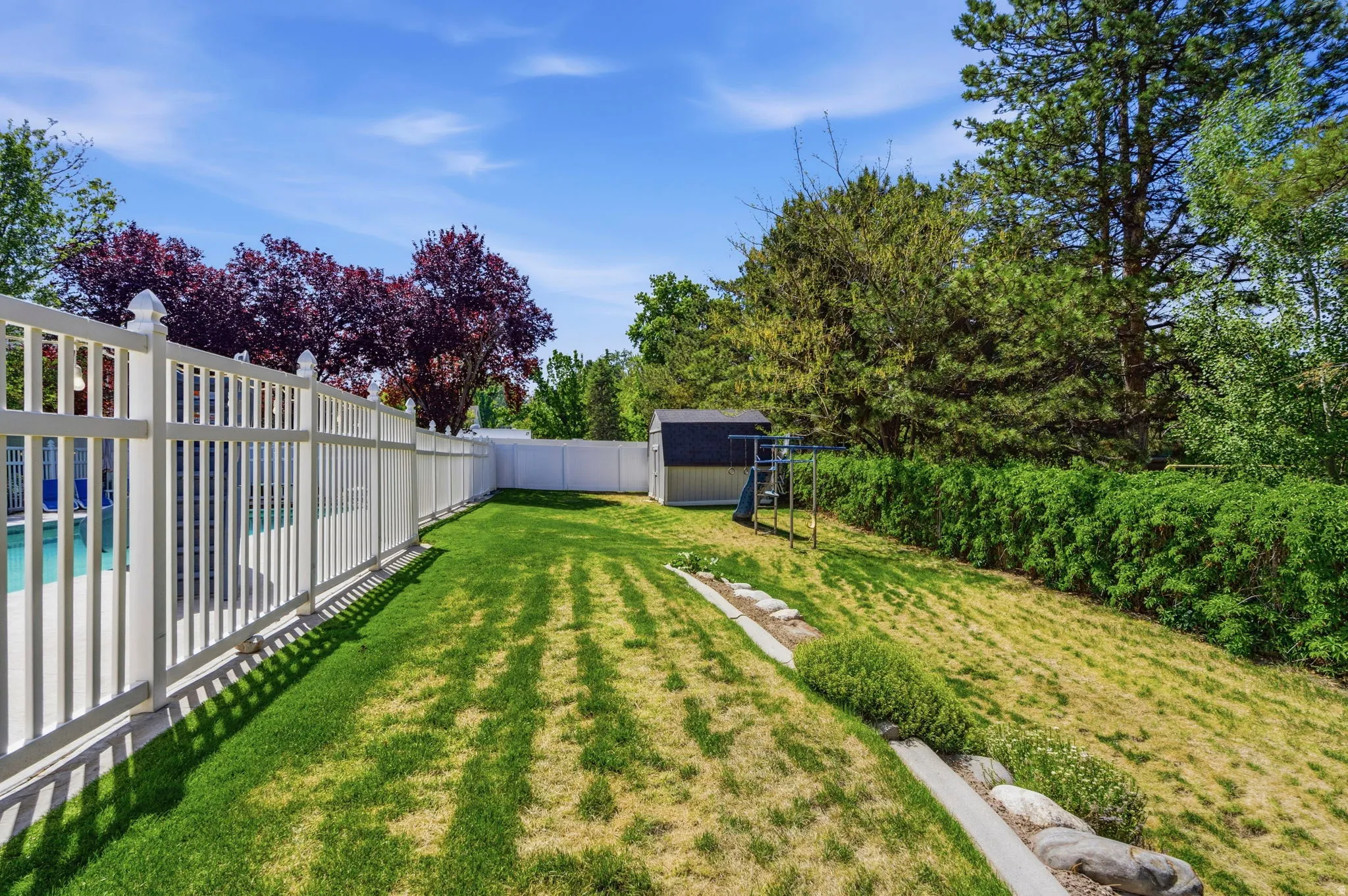 Expansive backyard featuring a white vinyl privacy fence, a detached shed, a sloped grassy area, mature trees, and a visible swimming pool portion
