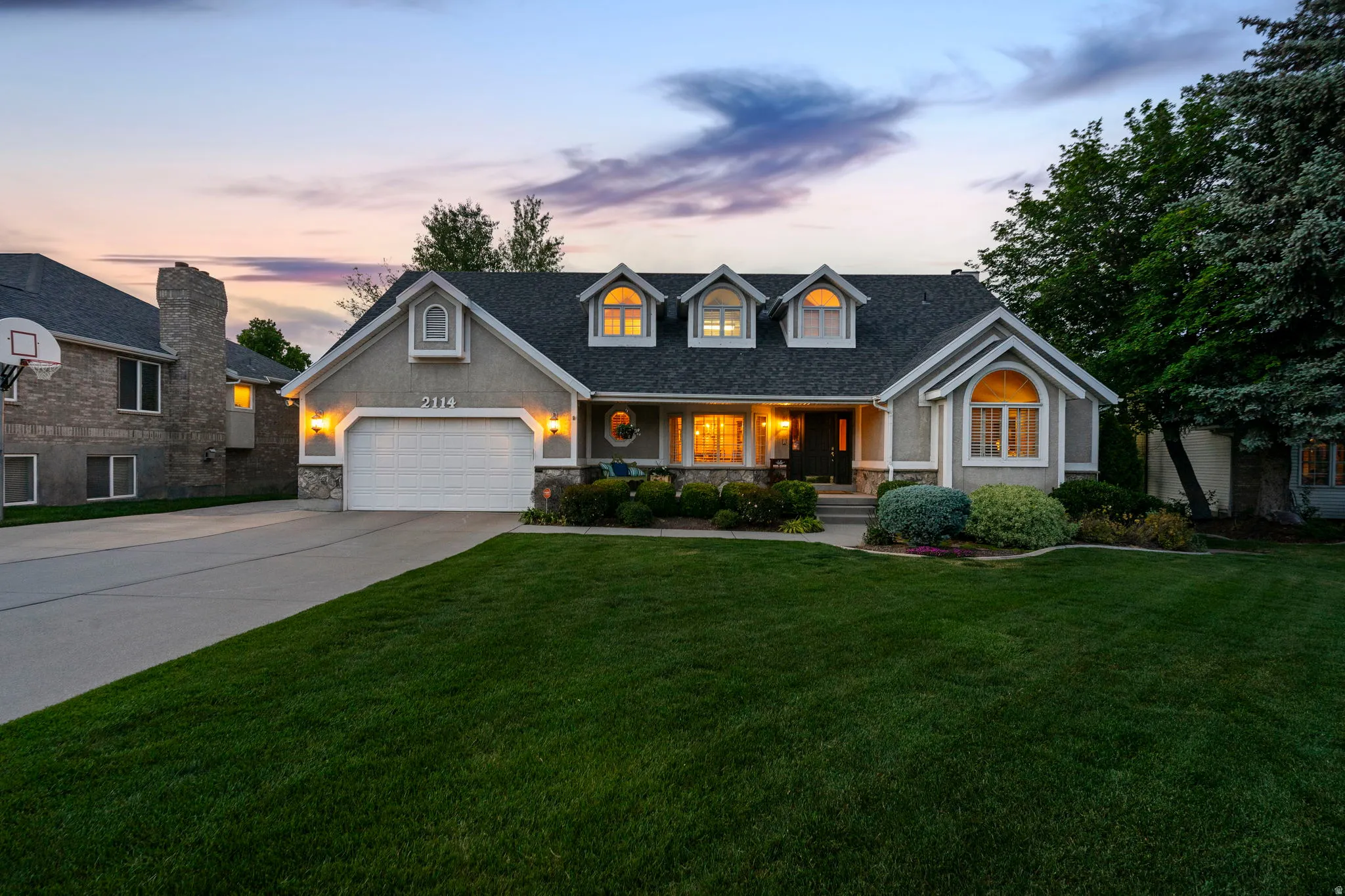 Traditional-style residence featuring a stucco and stone facade, dormer windows, a two-car garage, and mature landscaping