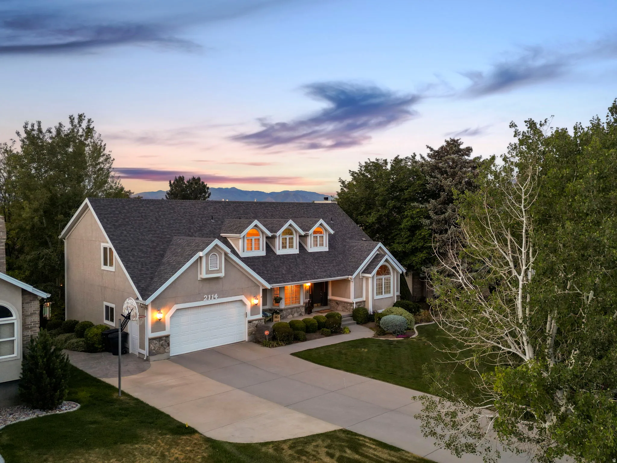 Traditional-style residence featuring a two-car garage, dormer windows, a concrete driveway, and mature landscaping