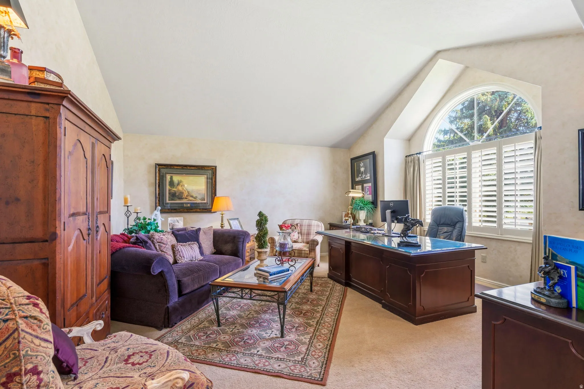 Vaulted ceiling room featuring a large arched window with plantation shutters, a built-in wood-tone desk, textured wall finish, and neutral carpet flooring
