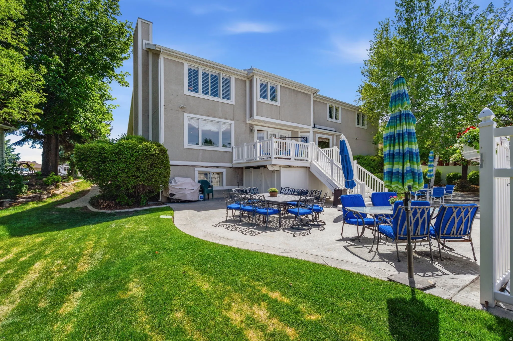 Expansive backyard featuring a large concrete patio, manicured lawn, multi-level deck with white railings, and mature trees