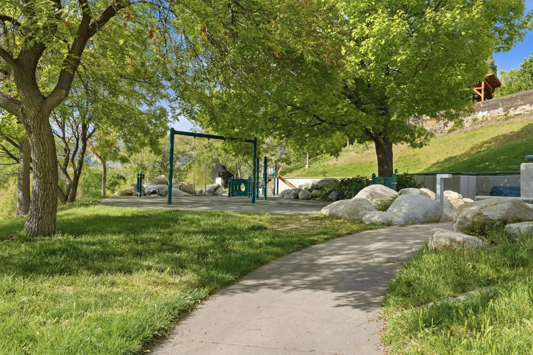 Serene park across the street featuring mature trees, a paved pathway, play equipment, natural rock landscaping, and a grassy hillside with incredible views