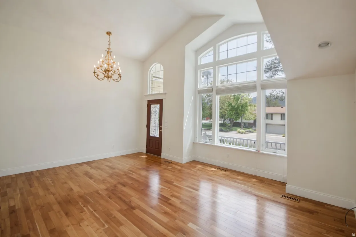 Formal living room/entry with vaulted ceiling great room featuring expansive picture windows, hardwood flooring, an arched transom window, a decorative front entry door, and a brass chandelier