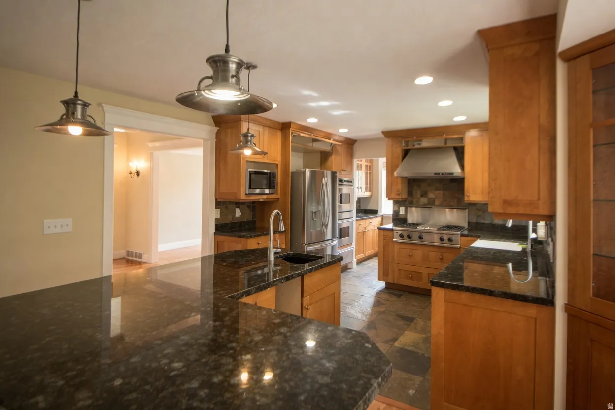 Kitchen featuring a spacious island with a dark countertop, stainless steel appliances, wood cabinetry, stone tile flooring, and pendant lighting
