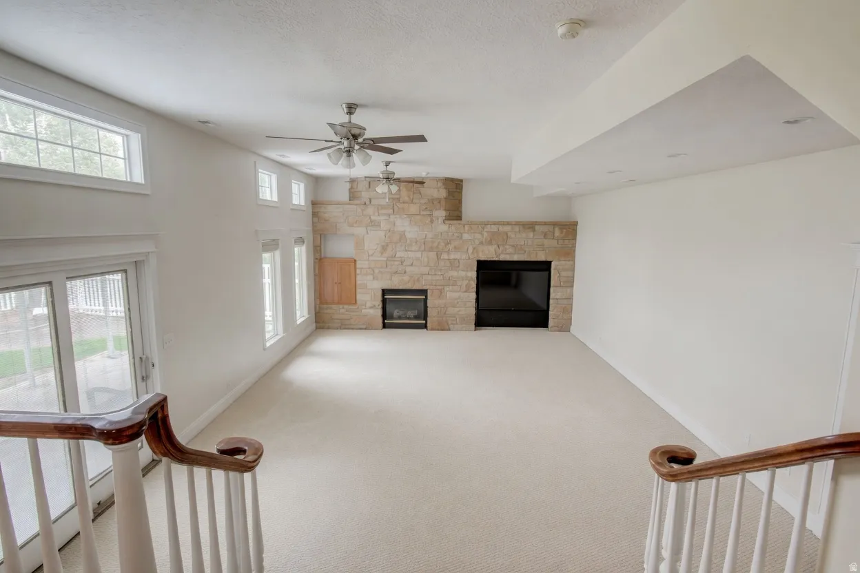 Expansive living space featuring a stone-clad fireplace surround, dual ceiling fans, multiple windows, and light-toned carpeting