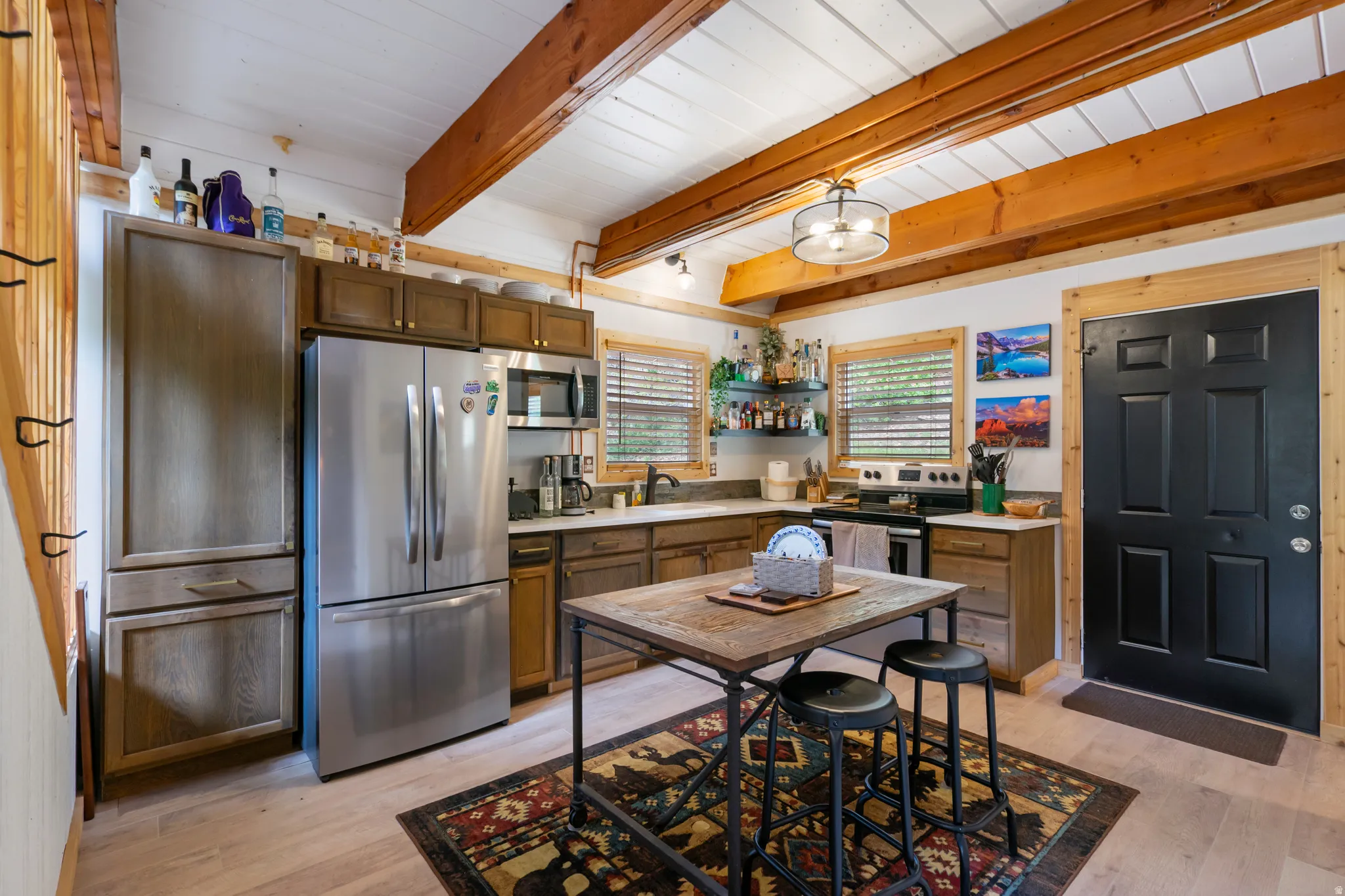 Kitchen featuring exposed wood beams, light wood-finish flooring, stainless steel appliances, dark wood cabinetry, and a black paneled door with natural wood trim