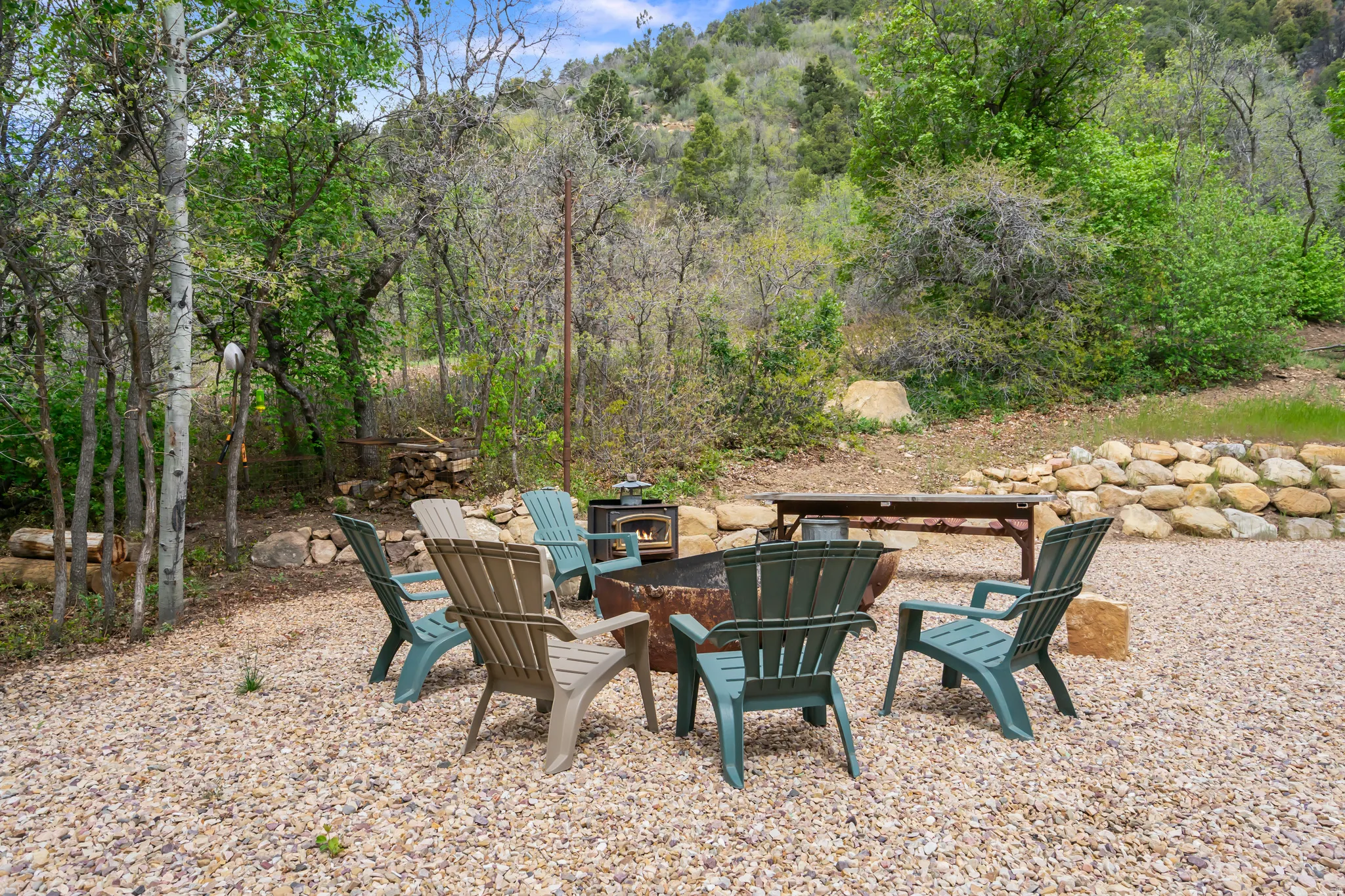Gravel patio area featuring a metal fire pit, surrounded by lush trees and natural landscaping