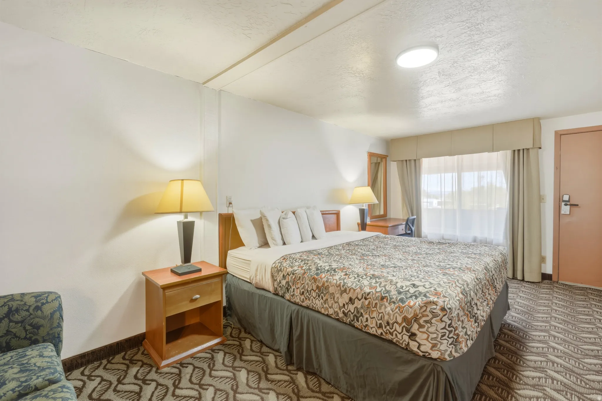 Bedroom featuring textured wall-to-wall carpeting, a large window with sheer and blackout draperies, and a built-in desk area