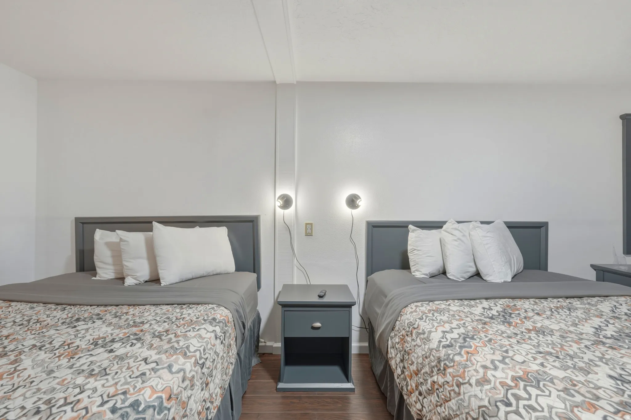 Bedroom featuring two beds with dark gray headboards, dark wood-finish flooring, white walls, and wall-mounted sconce lighting