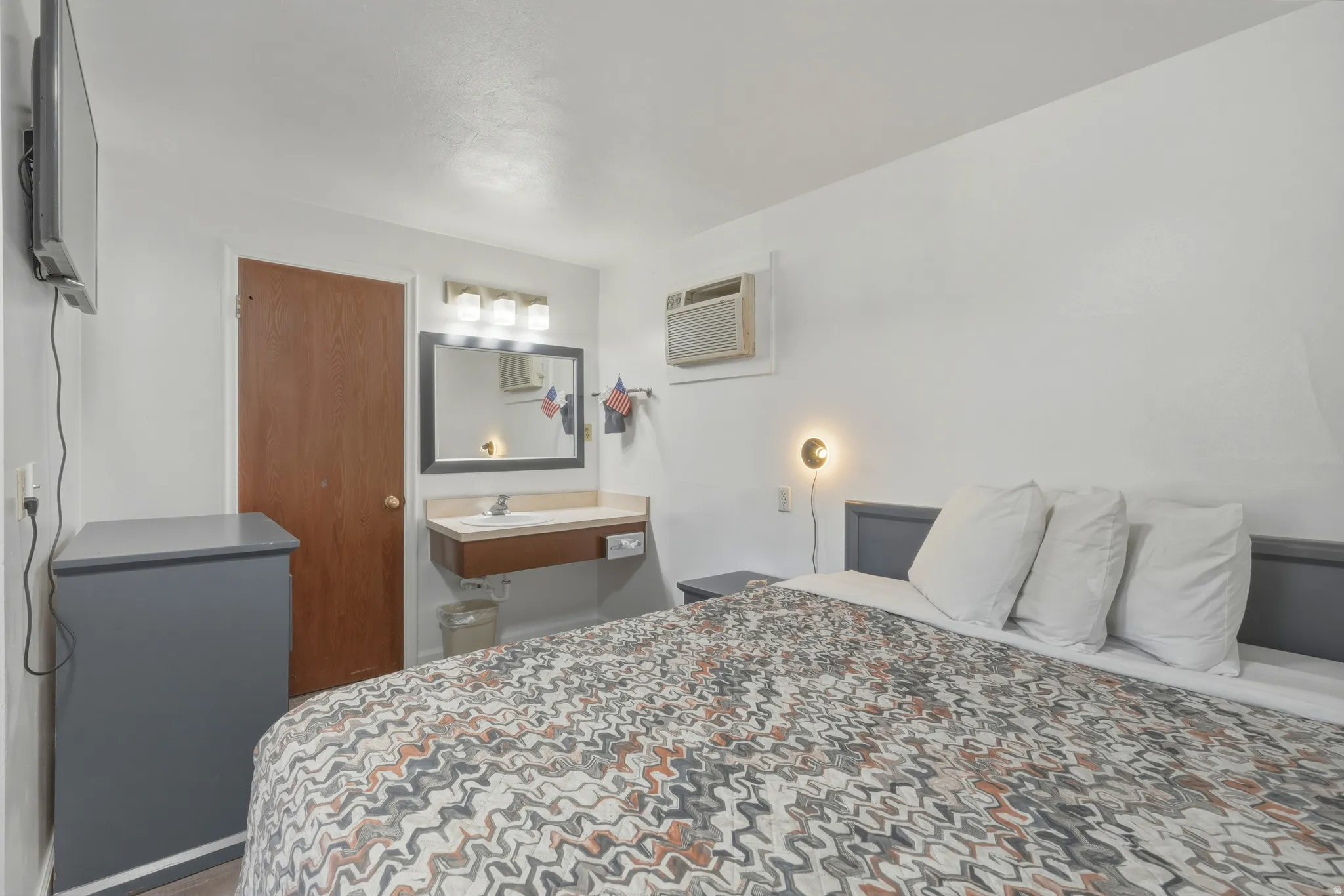 Bedroom featuring a wall-mounted television, a wood-finish door, a vanity sink with a framed mirror and three-light fixture, and an air conditioning unit