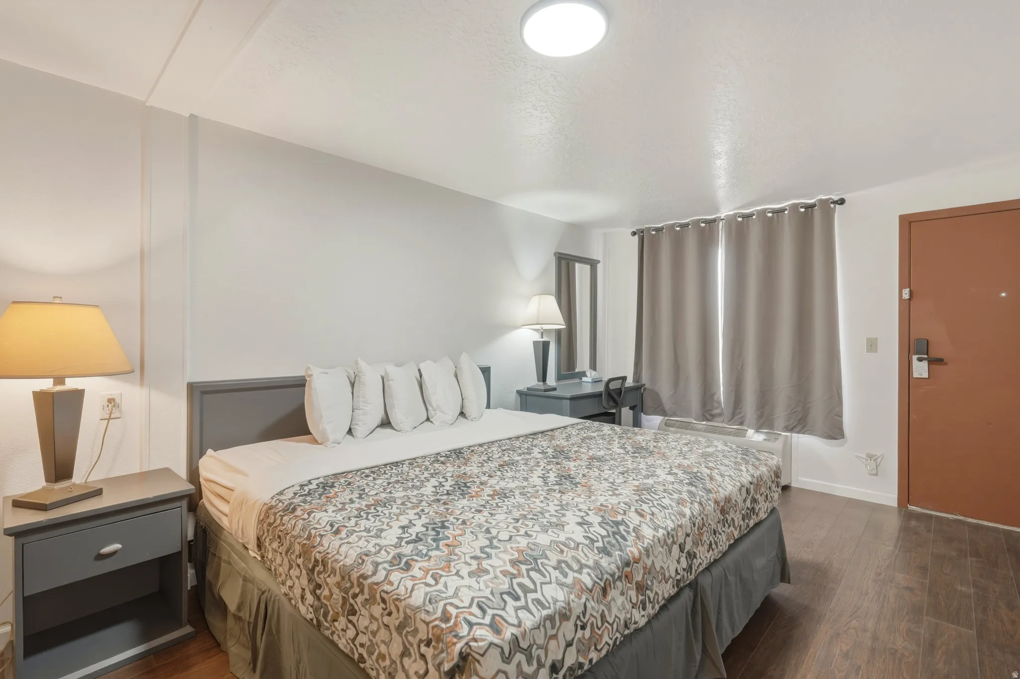 Bedroom featuring wood-finish flooring, a contemporary ceiling light fixture, a built-in wall-mounted AC unit, a desk area with a mirror, and window treatments