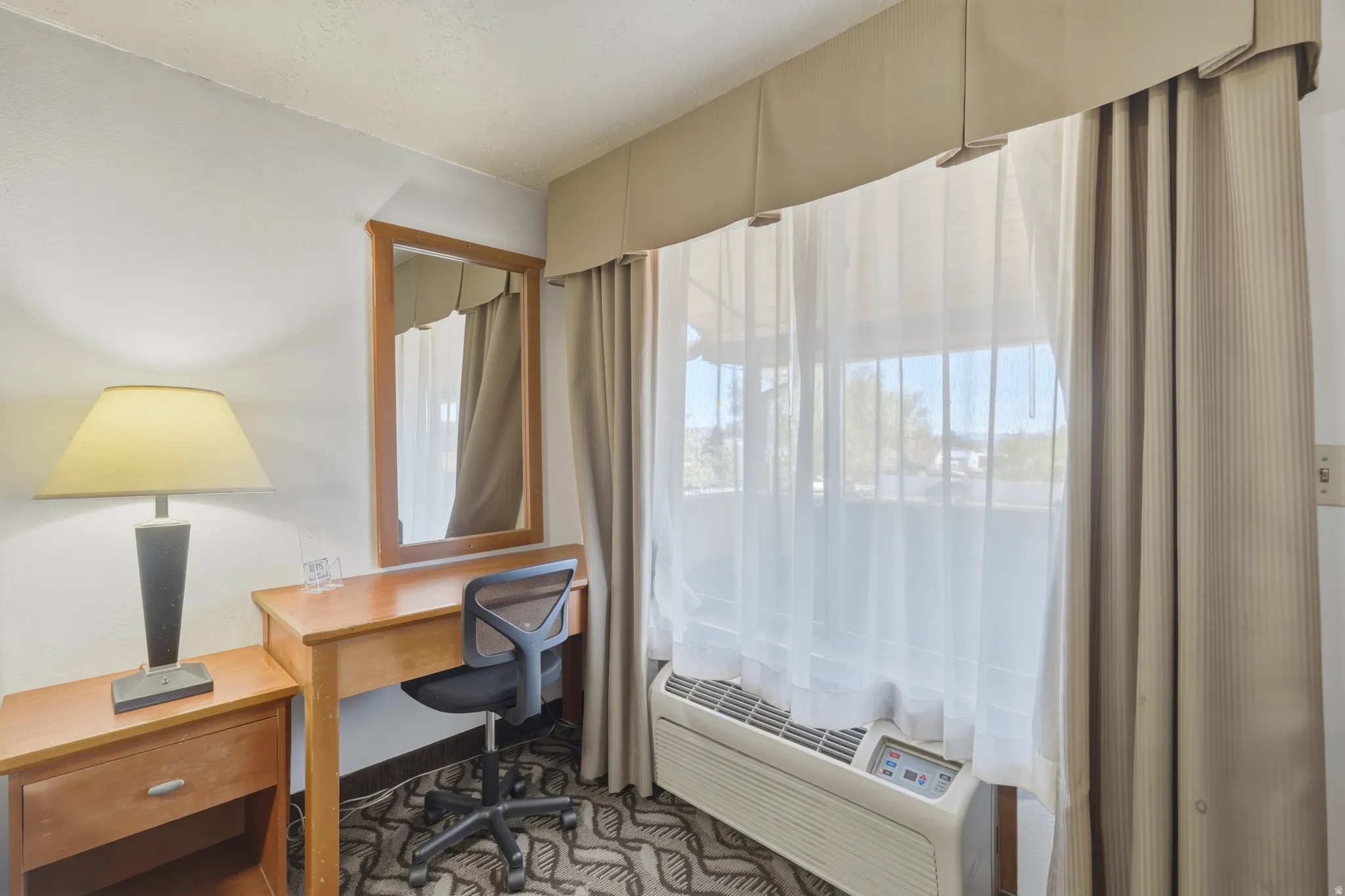 Window featuring sheer white curtains and beige drapes, wood-finish desk with a matching mirror, and patterned carpet flooring