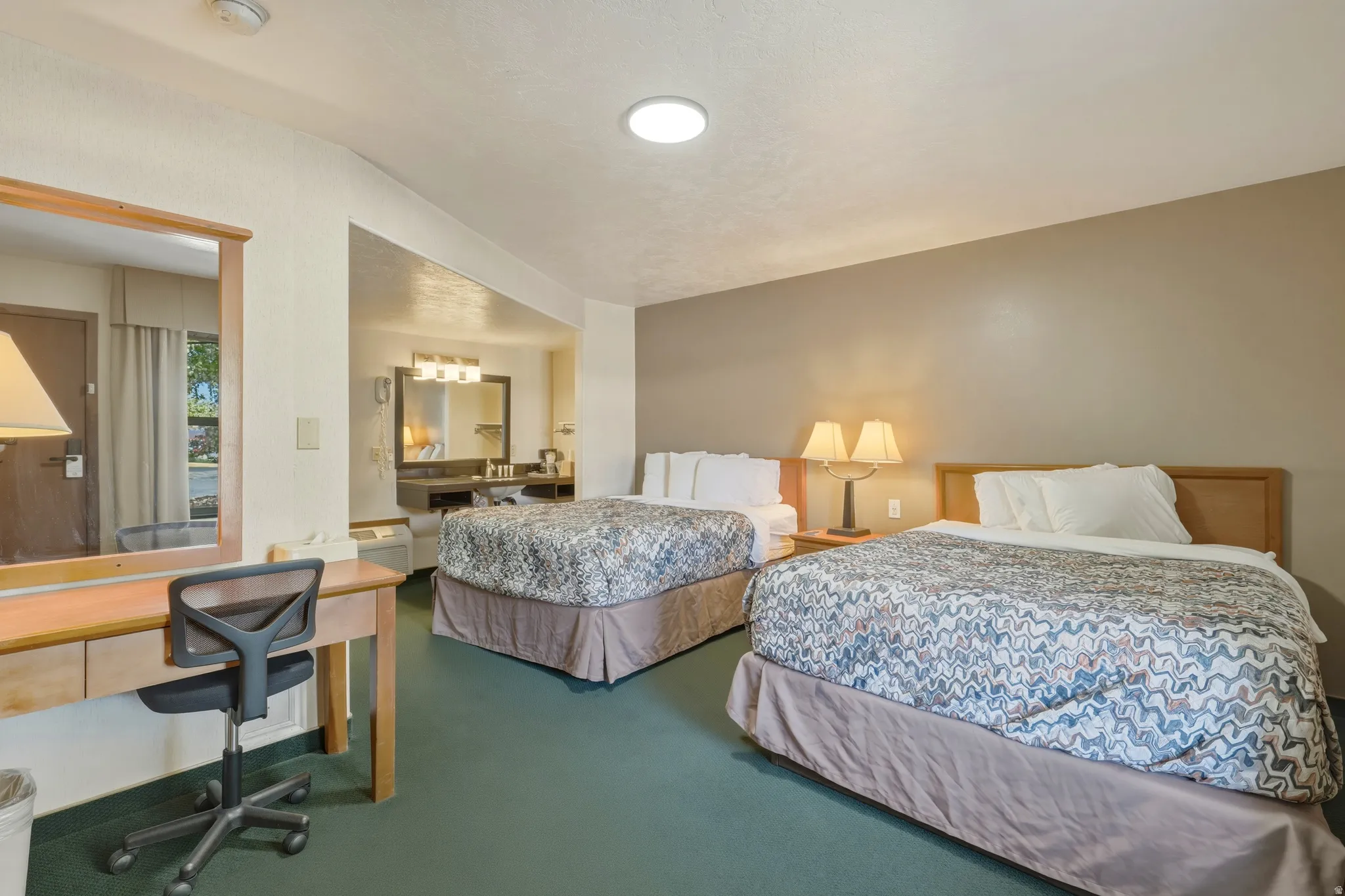 Bedroom featuring two beds with patterned bedding, a desk with a mirror, and a vanity area with a mirror and integrated lighting