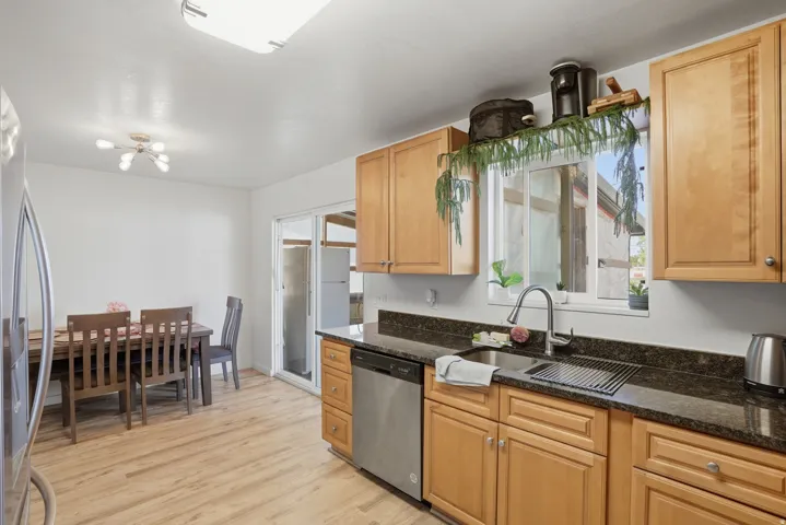 Kitchen featuring light wood-finish cabinetry, dark countertops, a stainless steel sink with gooseneck faucet, a stainless steel dishwasher, and wood-finish flooring