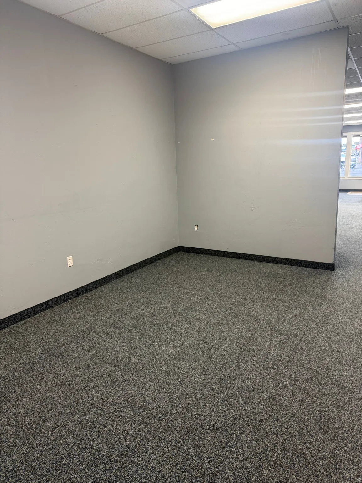 Spacious interior room with light gray walls, dark gray carpet, black baseboards, and a white grid ceiling featuring flush-mounted lighting