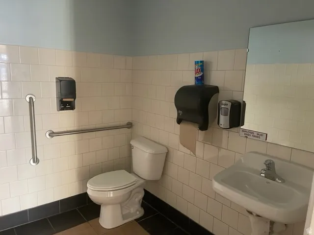 Restroom featuring white subway tile walls, a wall-mounted sink with chrome faucet, and a toilet with grab bars