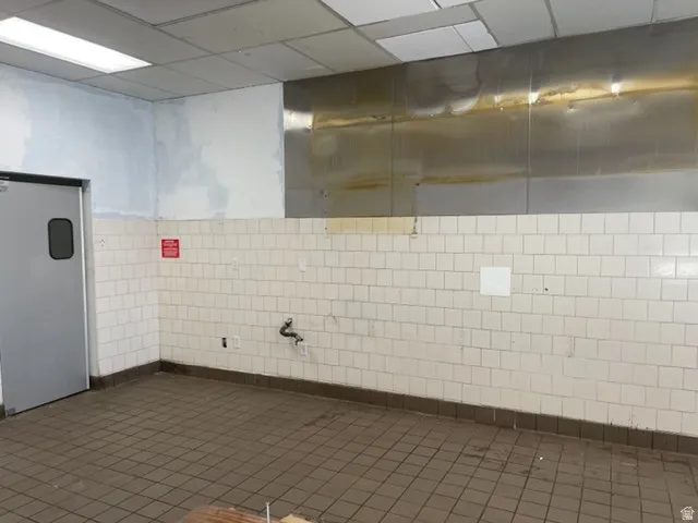 Commercial interior featuring white square wall tiles with a dark base trim, grey square floor tiles, and a drop ceiling with integrated fluorescent lighting