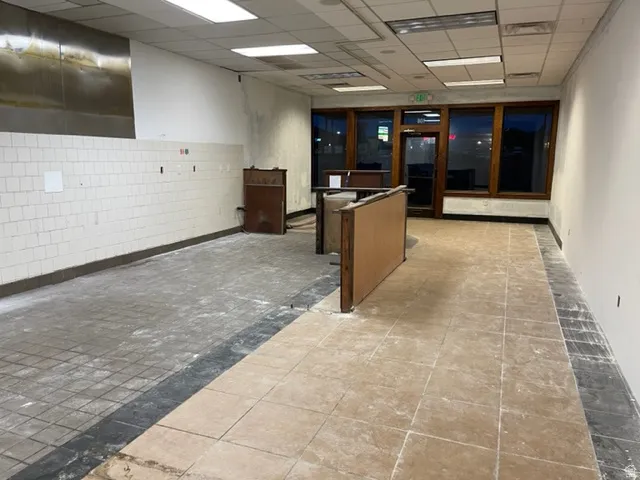 Spacious commercial interior featuring a mix of square tile flooring, exposed ceiling grid with recessed lighting, and a partial wall of white square tiles