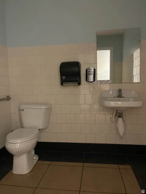 Restroom featuring white square wall tiling, a white wall-mounted sink, a matching white toilet, and contrasting dark baseboard tiling