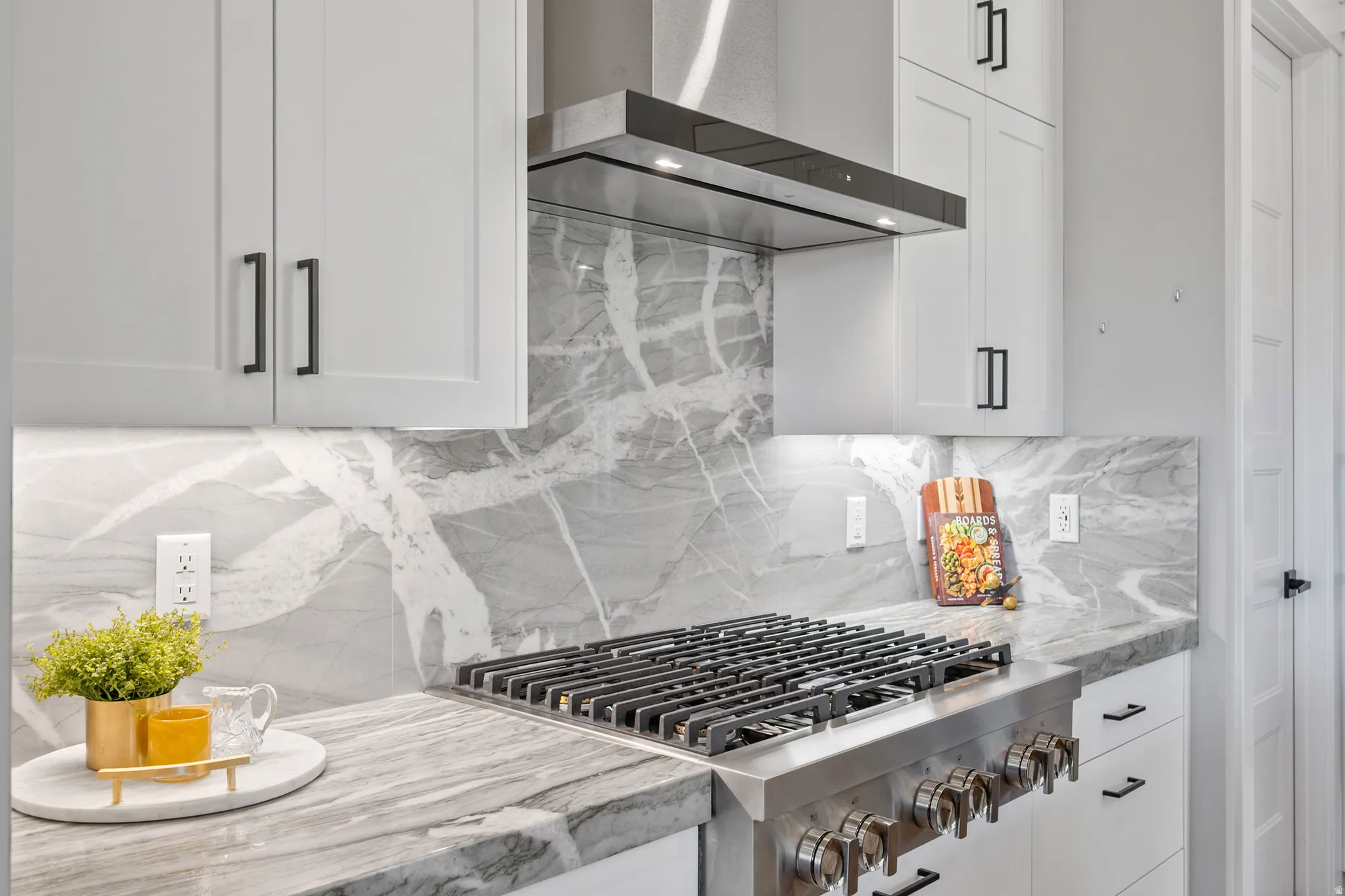 Gourmet kitchen featuring a stainless steel range with griddle, coordinating range hood, full-height veined backsplash, stone countertops, and white shaker-style cabinetry with matte black hardware
