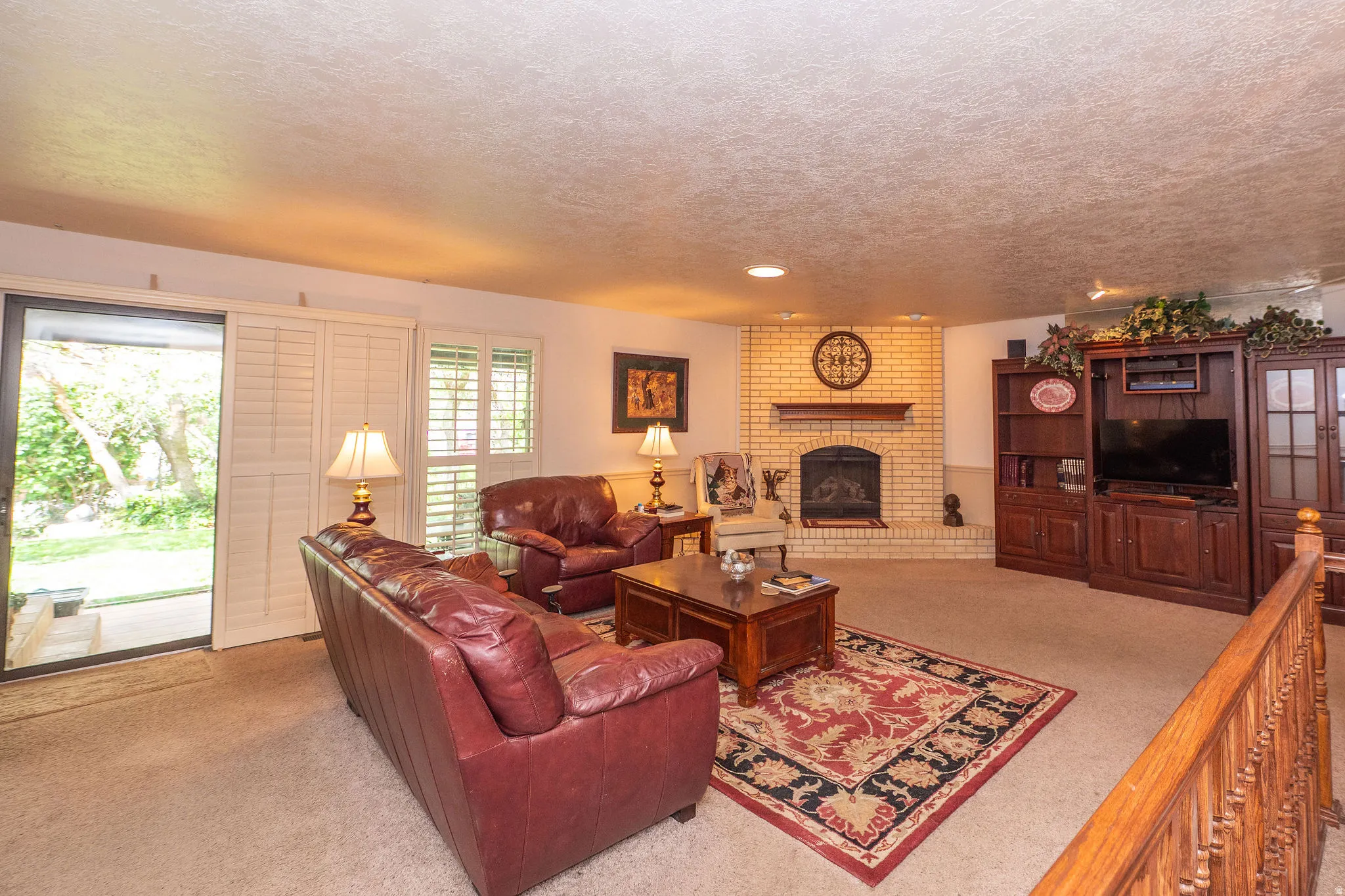 Spacious living area featuring a light-colored brick fireplace with a wood mantle, neutral carpet flooring, and a sliding glass door providing outdoor access