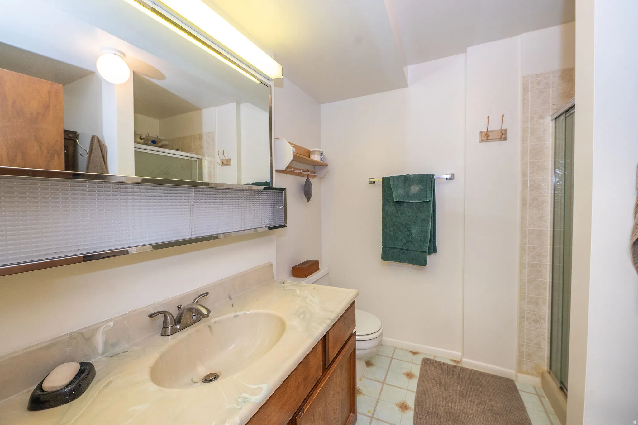 Bathroom featuring a wood-finish vanity with an integrated sink and light-toned countertop, a large mirror with integrated lighting, a toilet, a shower stall with tiled walls, and patterned floor tiling