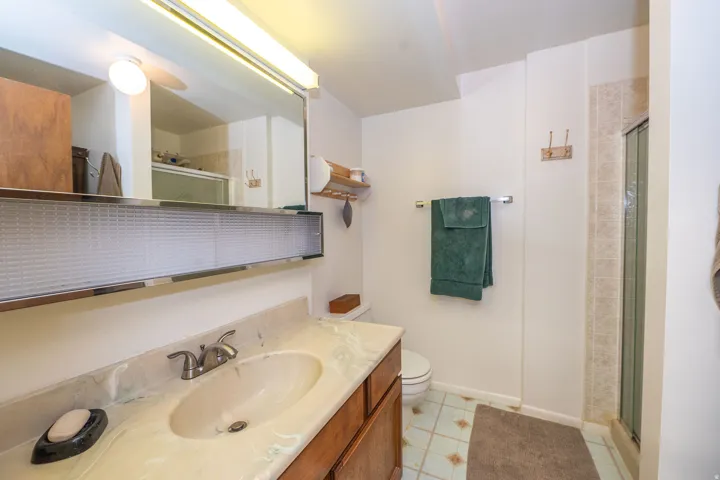 Bathroom featuring a wood-finish vanity with an integrated sink and light-toned countertop, a large mirror with integrated lighting, a toilet, a shower stall with tiled walls, and patterned floor tiling