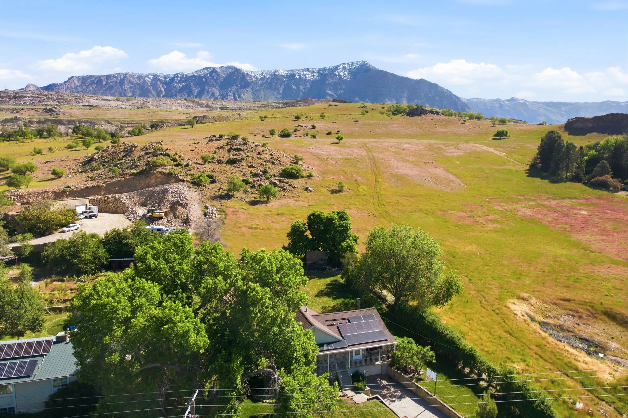Expansive property featuring a residence with solar panels, mature trees, and a concrete driveway, set against a backdrop of rolling hills and distant snow-capped mountains
