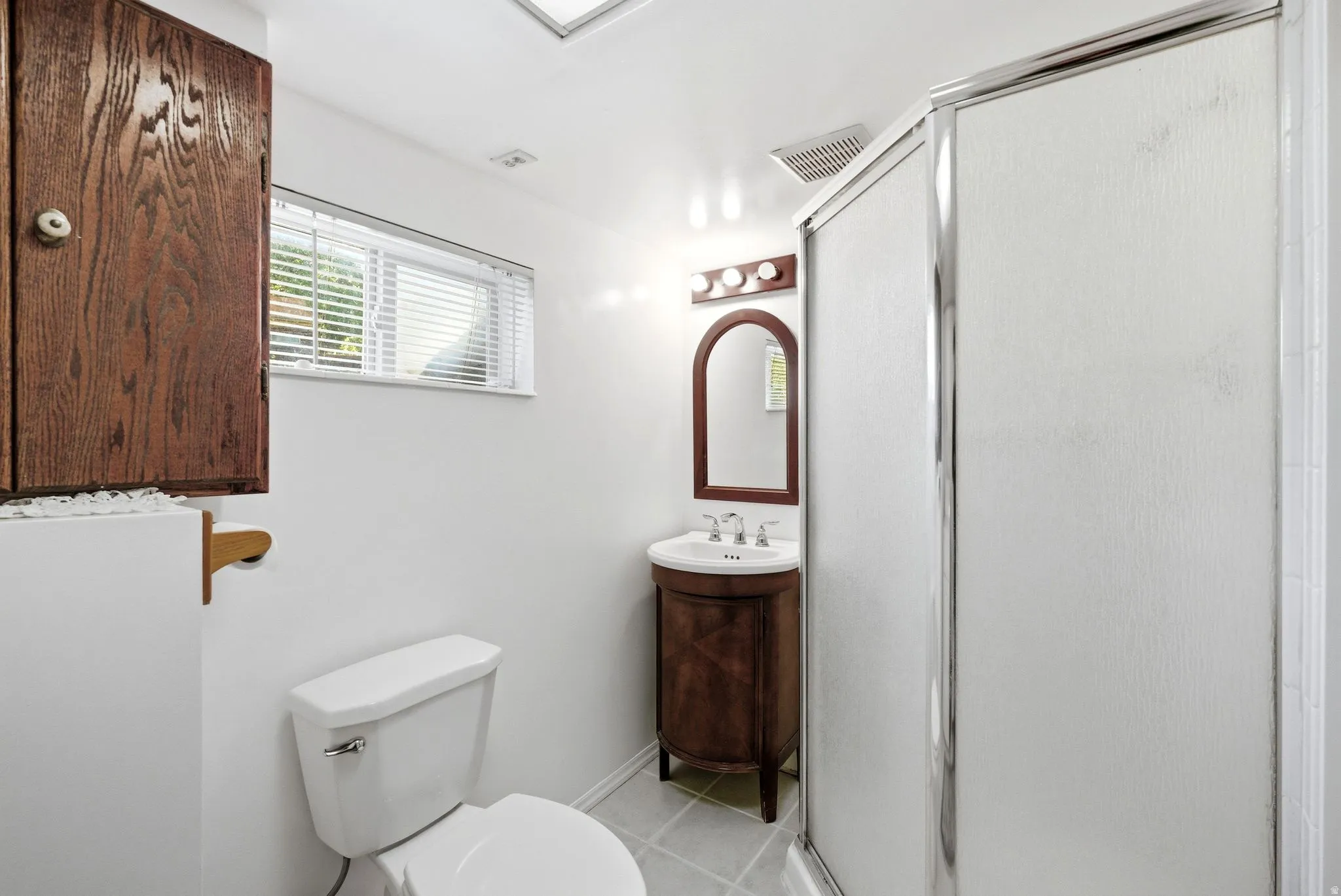 Bathroom featuring a glass-enclosed shower, a wood-finish vanity with an arched mirror, an overhead light fixture, a window with blinds, and white square floor tiles