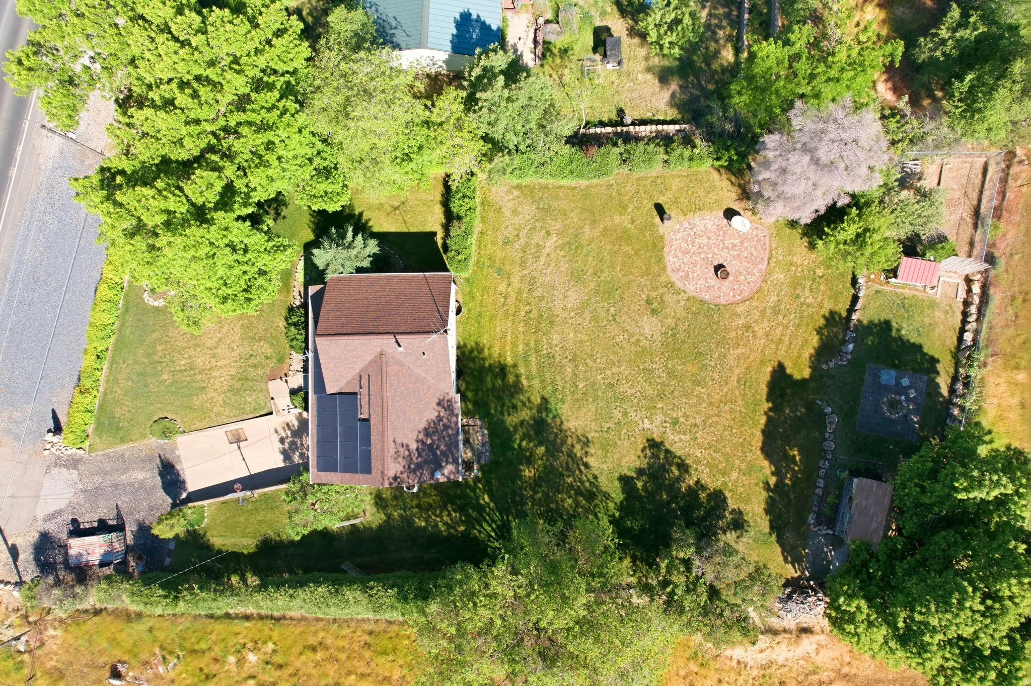 Expansive property featuring a brown shingle roof, solar panels, a paved driveway with a basketball hoop, a large grassy area, and a circular wood chip patio with a central fire pit