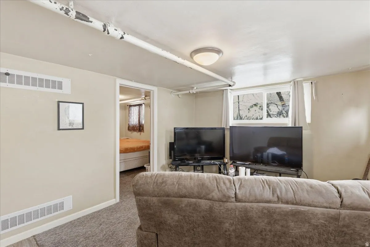 Carpeted room featuring a white ceiling with exposed piping, a single flush-mount light fixture, and a window providing natural light