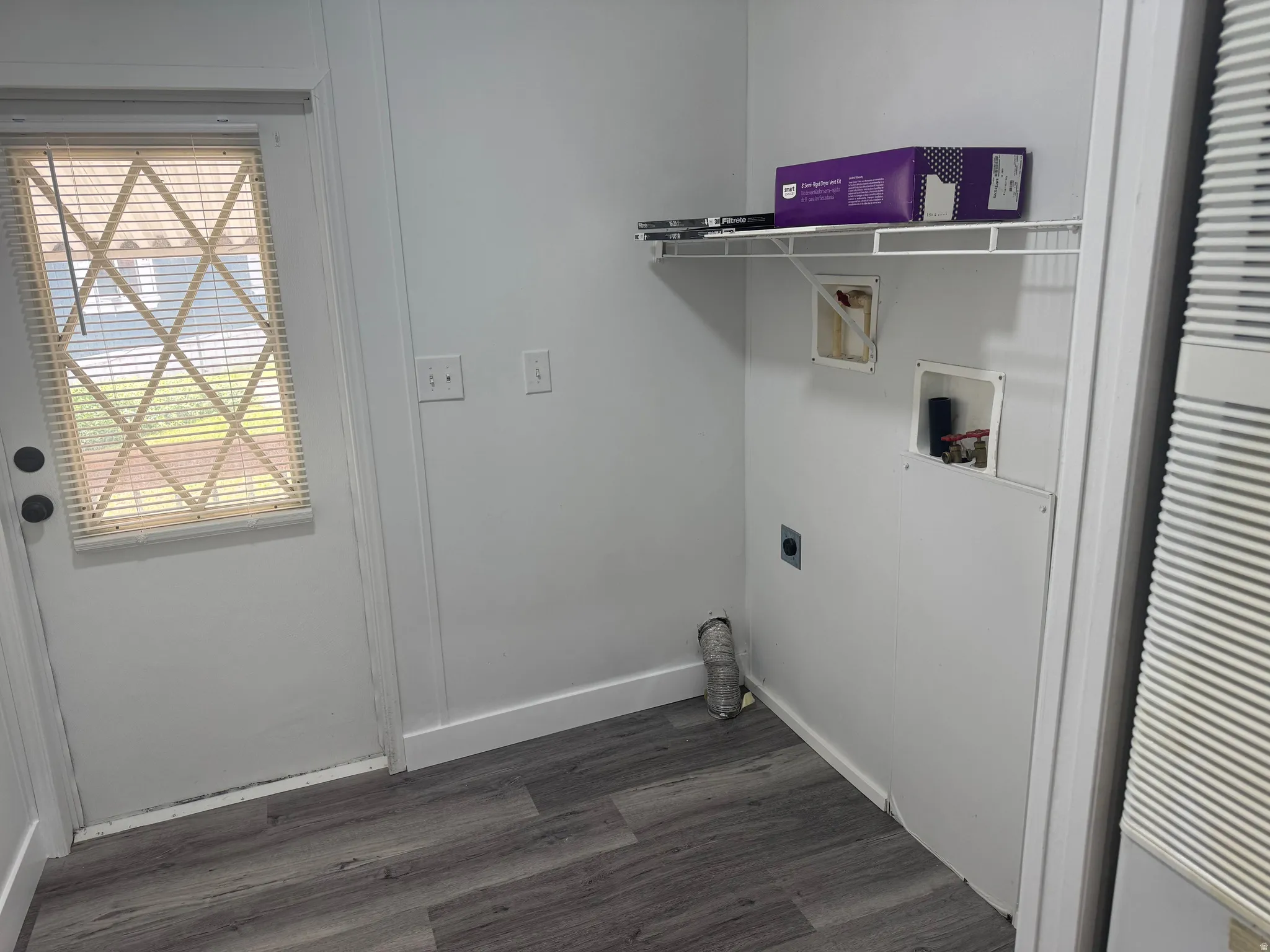 Laundry area featuring wood-finish flooring, a white utility door with integrated blinds, and a wall-mounted wire shelf