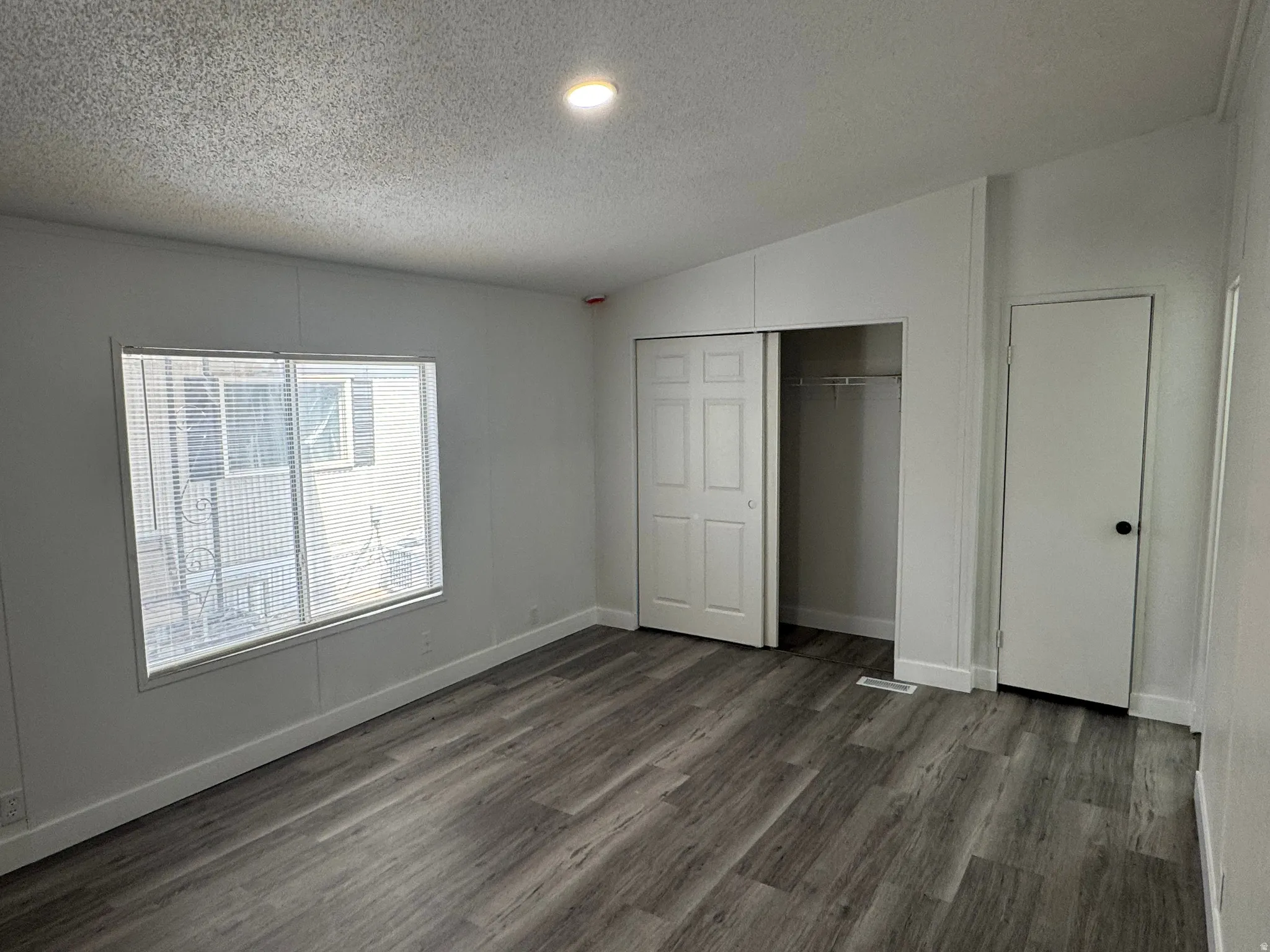Spacious room featuring wood-finish flooring, a large window with blinds, a built-in closet with bi-fold doors, a recessed ceiling light, and white interior doors