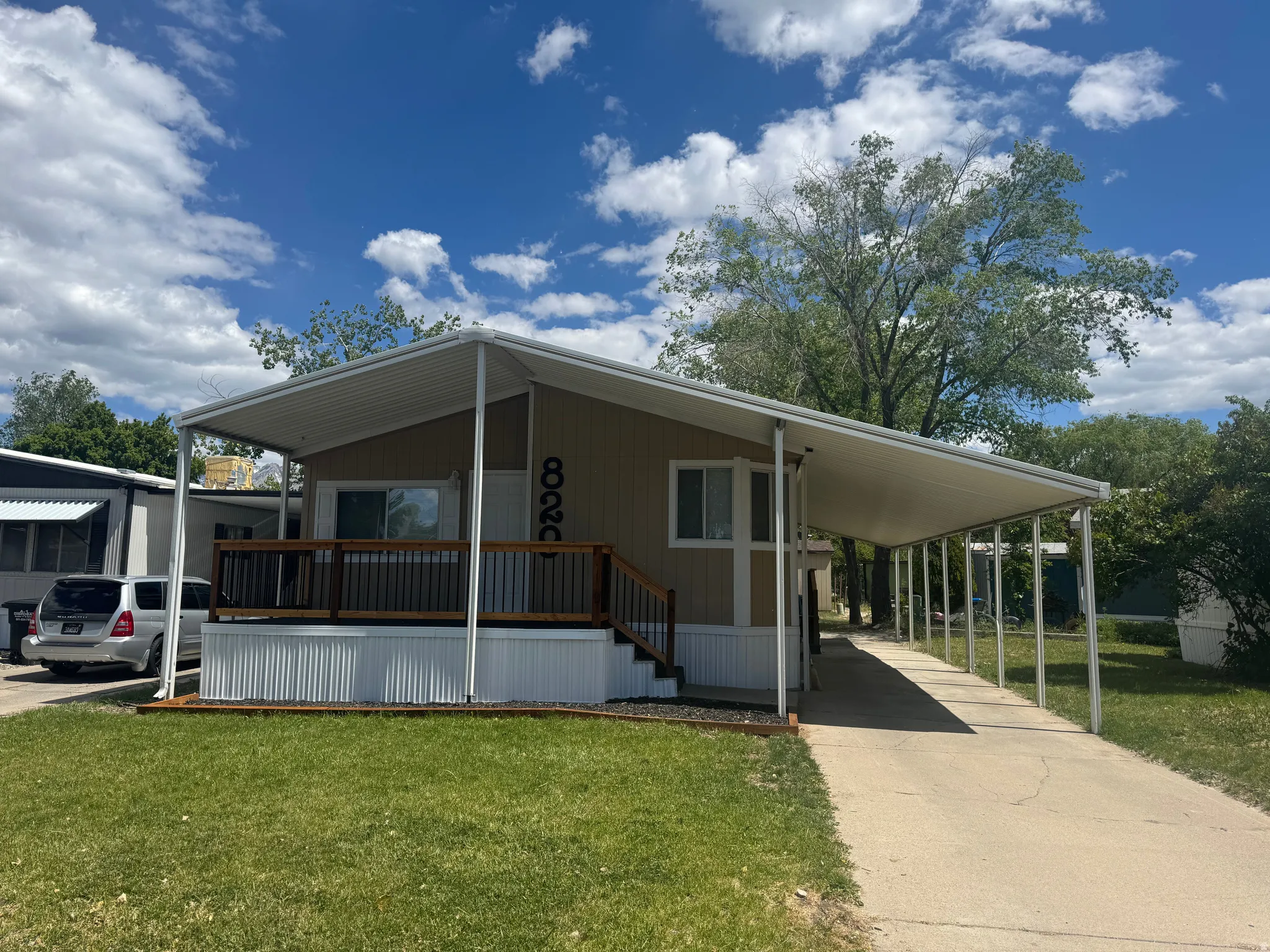 Single-story manufactured home featuring a tan siding exterior, a covered front porch with wood-finish decking and dark railings, and a concrete driveway leading to a car port