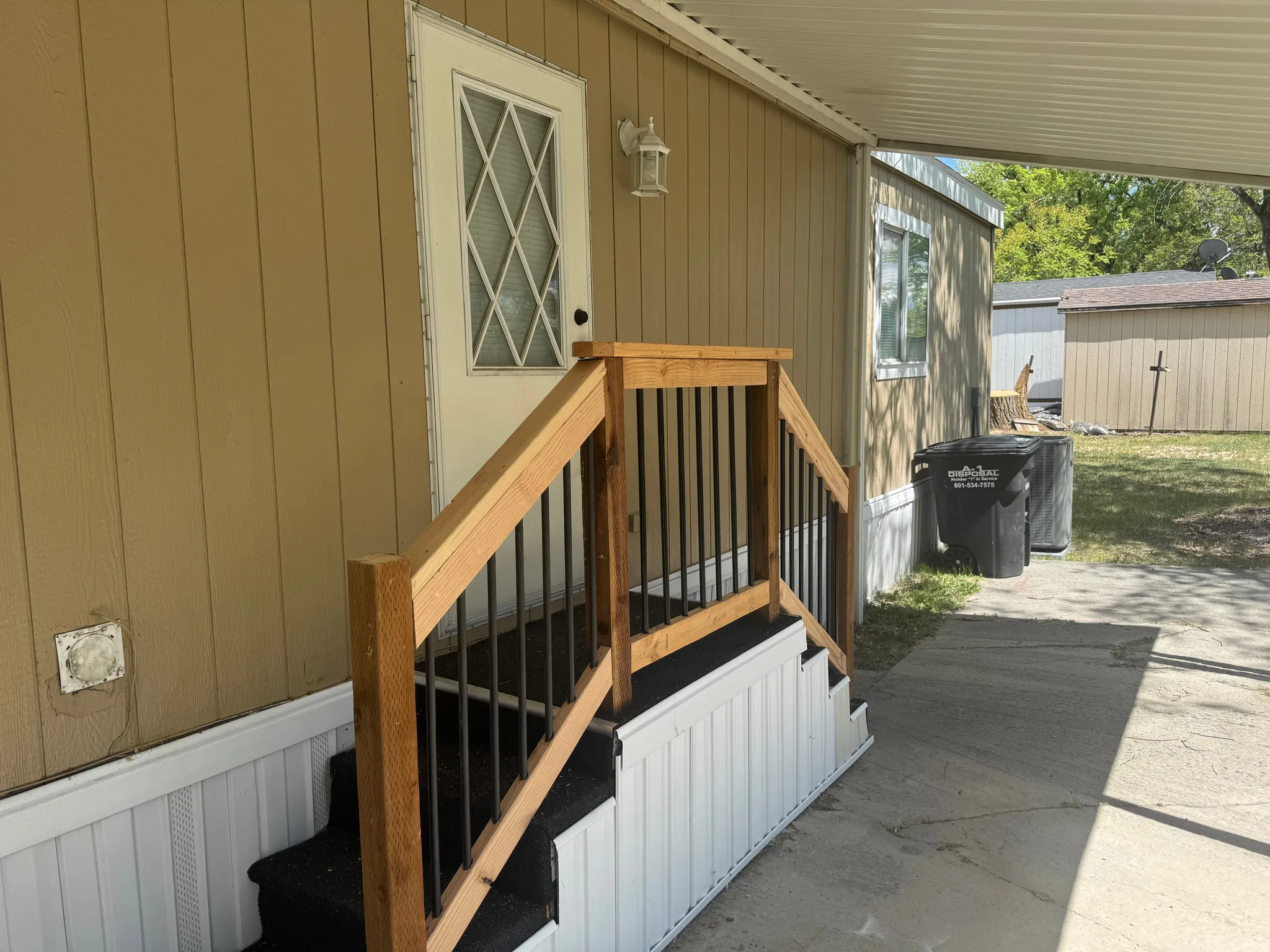 Exterior entry featuring a white door with diamond-patterned glass, natural wood handrail with dark balusters, white-skirted steps with dark tread, and tan vertical siding