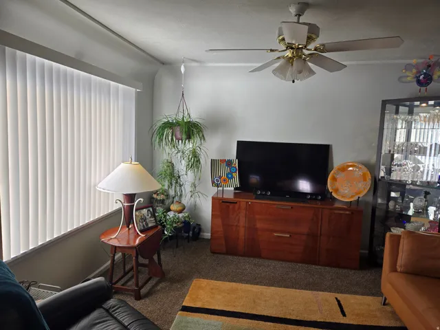 Living area featuring neutral wall paint, carpet flooring, a ceiling fan with integrated lighting, and vertical window blinds