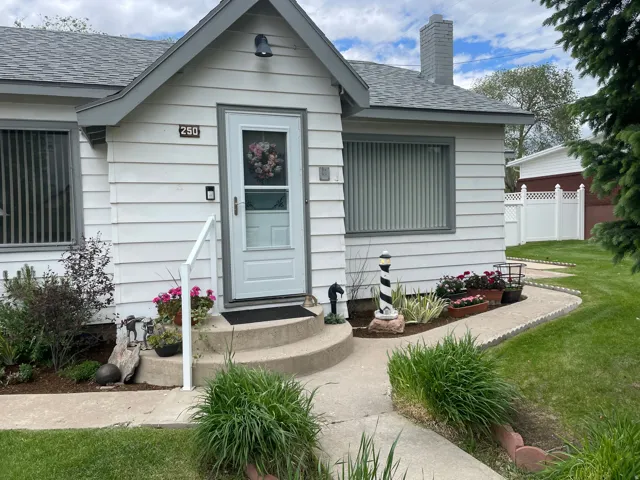 White siding exterior with grey trim accents, a grey shingled roof, a paved walkway, established shrubbery, and a white fence