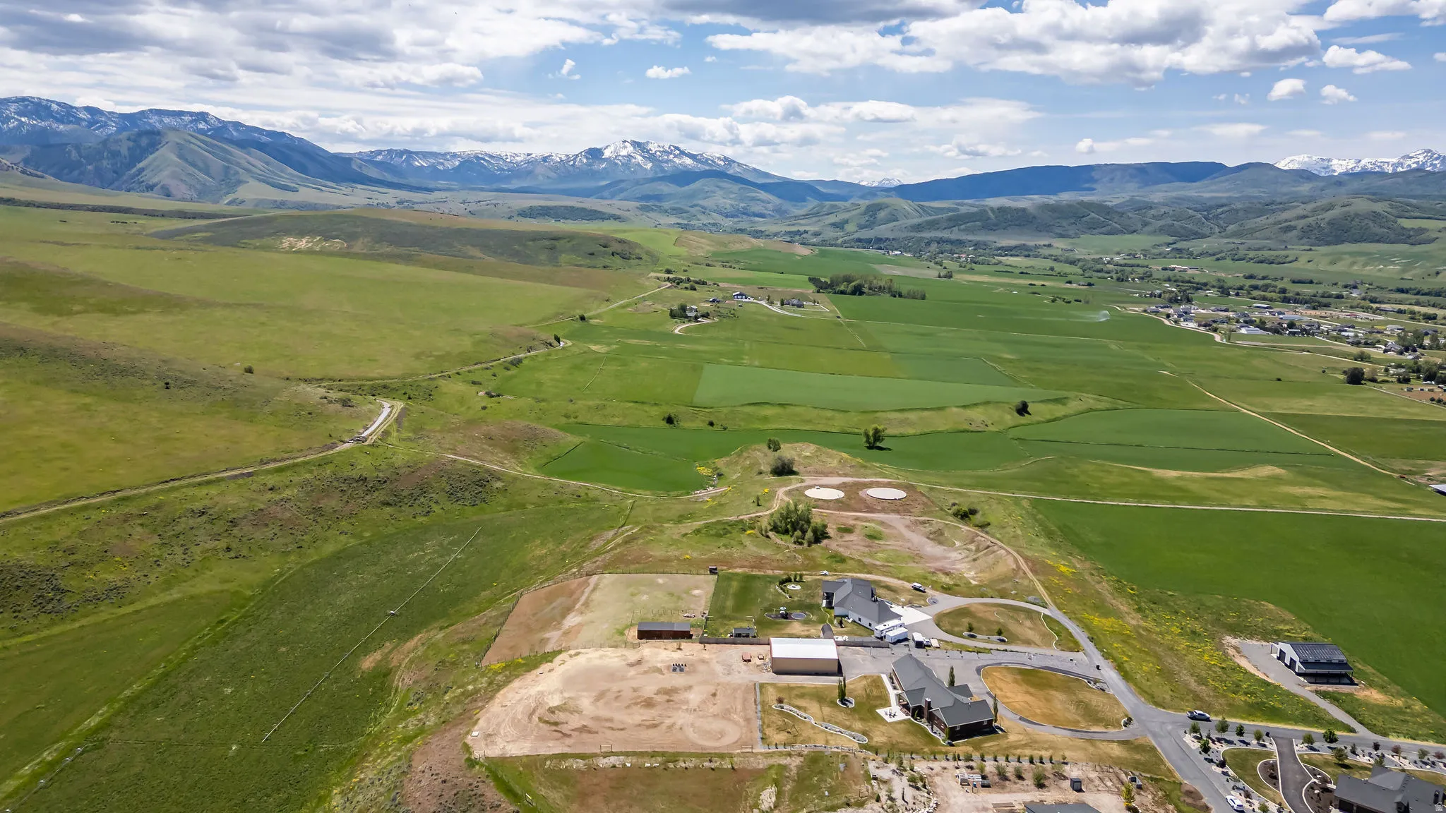 Expansive rural landscape featuring green fields, distant snow-capped mountains, and a clear sky with scattered clouds