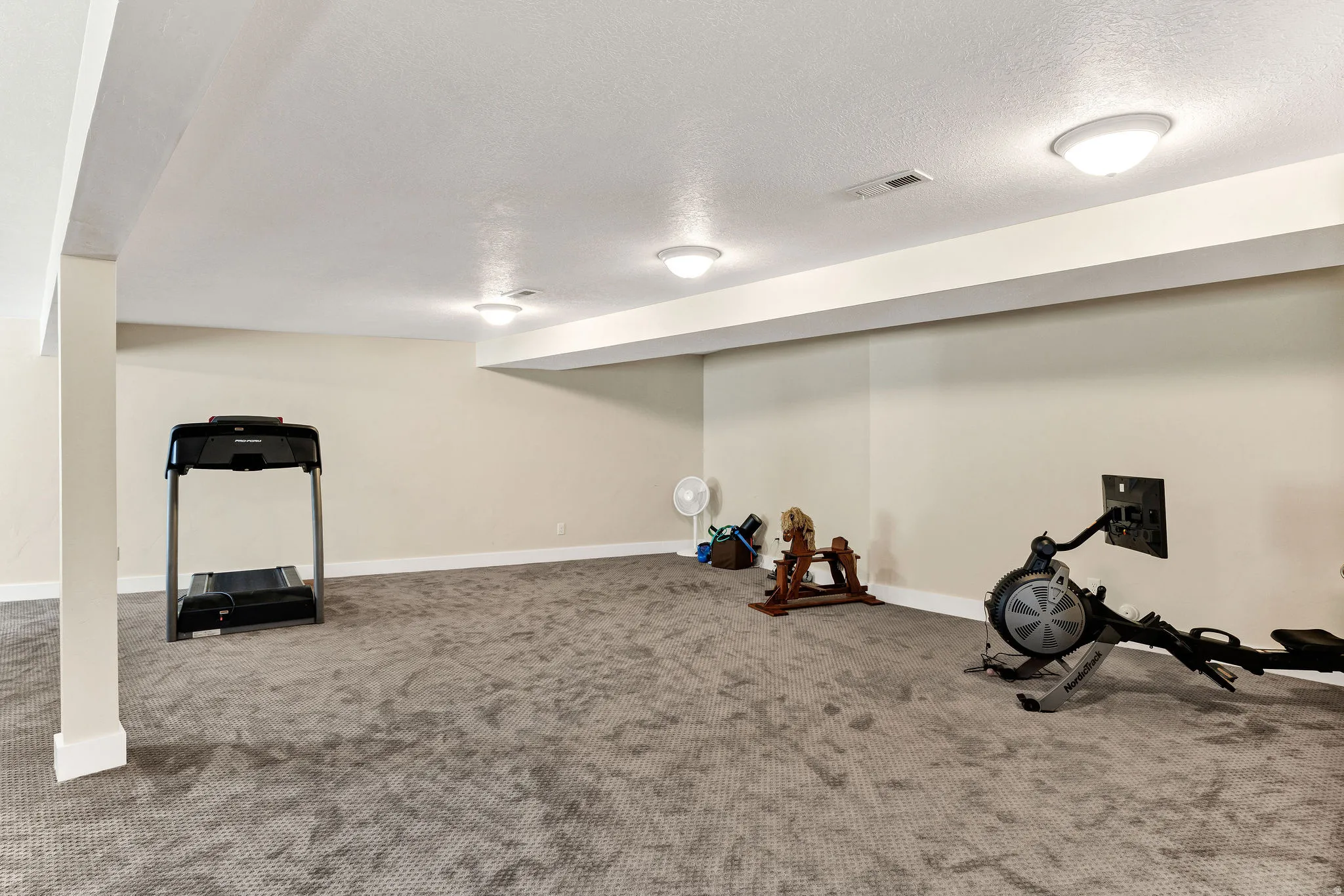 Spacious lower-level room featuring light neutral wall tones, gray carpeting, recessed ceiling lighting, and exposed structural beams