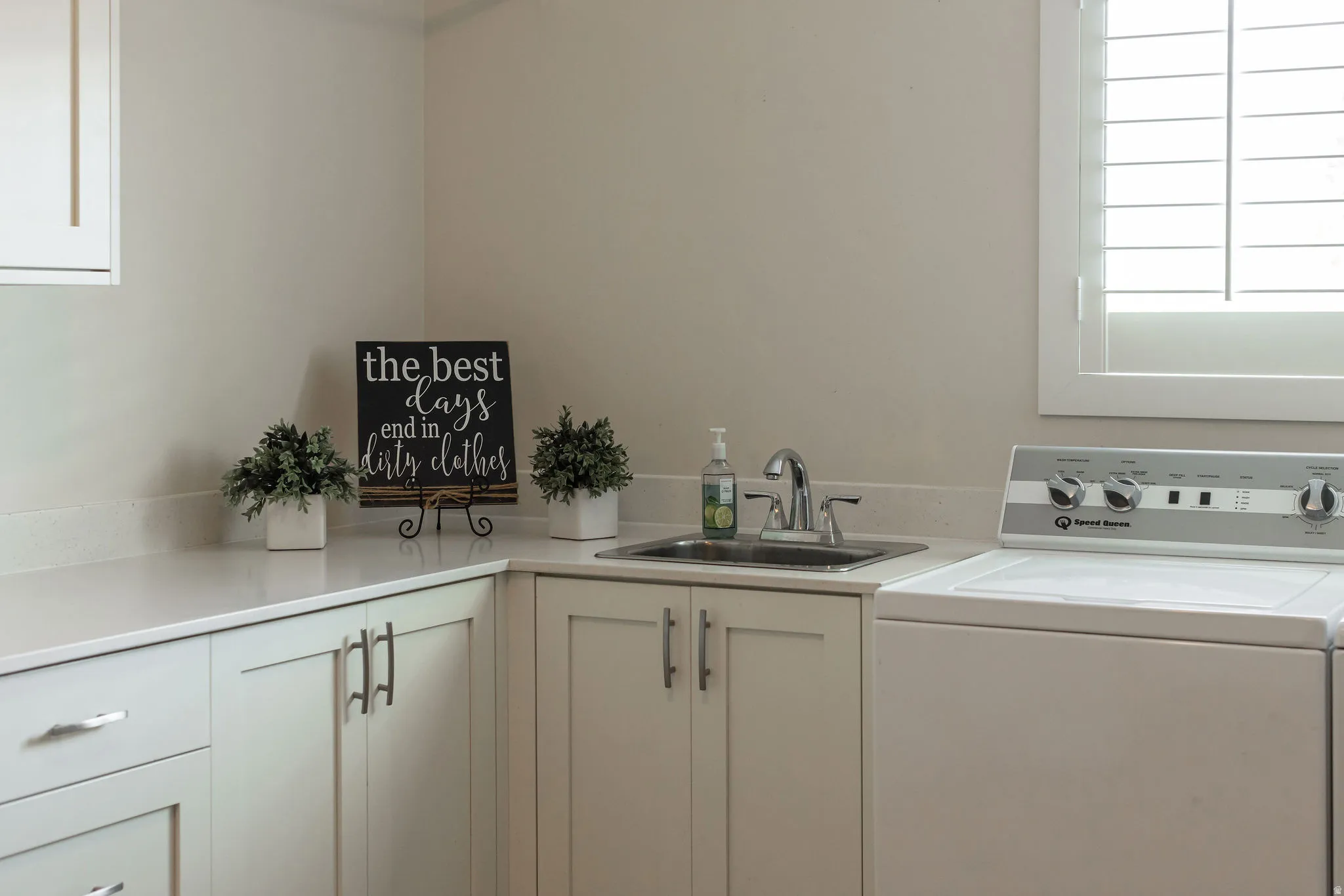 Laundry area featuring white cabinetry, light-toned countertops, a stainless steel sink with a chrome faucet, and a window with white plantation shutters