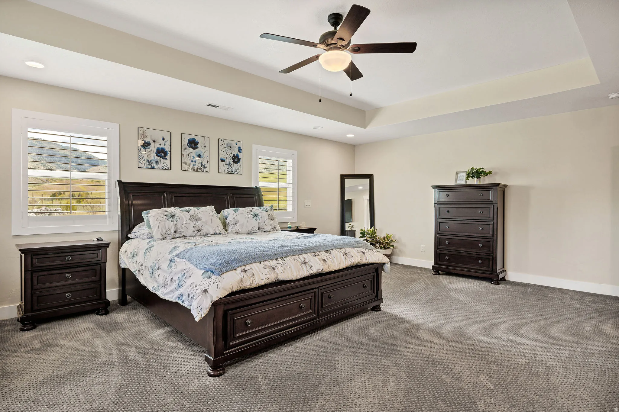 Spacious bedroom featuring neutral wall tones, tray ceiling, and ceiling fan with integrated lighting
