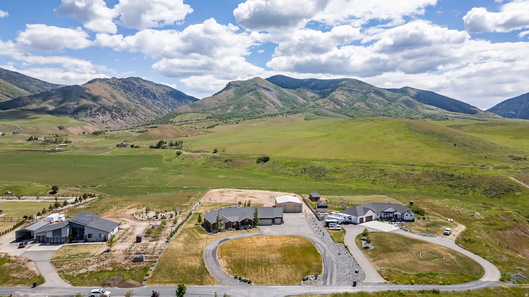 Expansive property featuring a modern ranch-style residence, a circular driveway, an attached multi-car garage, and a detached outbuilding, all set against a backdrop of rolling green hills and distant mountains