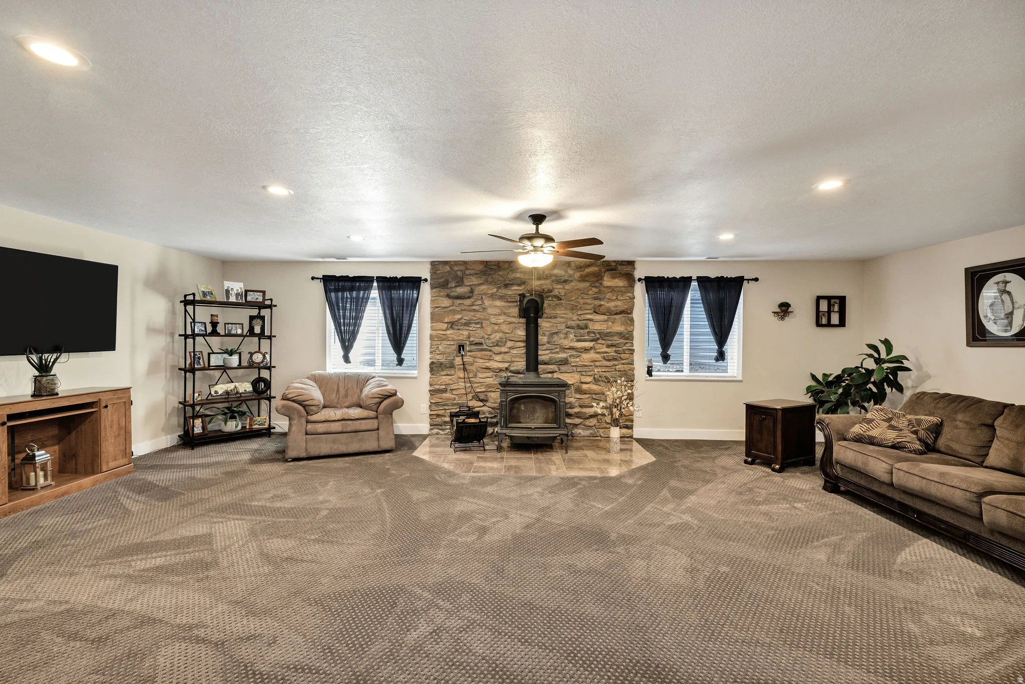 Spacious room featuring a stone-clad fireplace with a wood-burning stove, two windows, recessed lighting, and a ceiling fan
