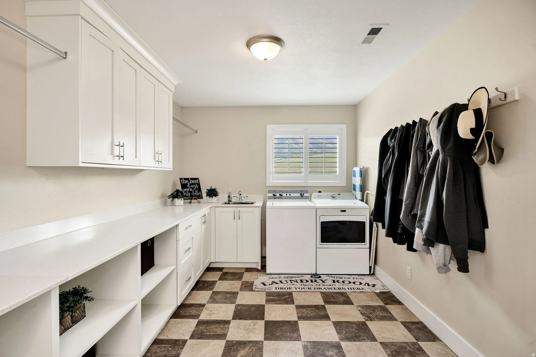 Spacious utility room featuring white cabinetry, extensive counter space, checkerboard flooring, a dedicated sink, and a window with plantation shutters