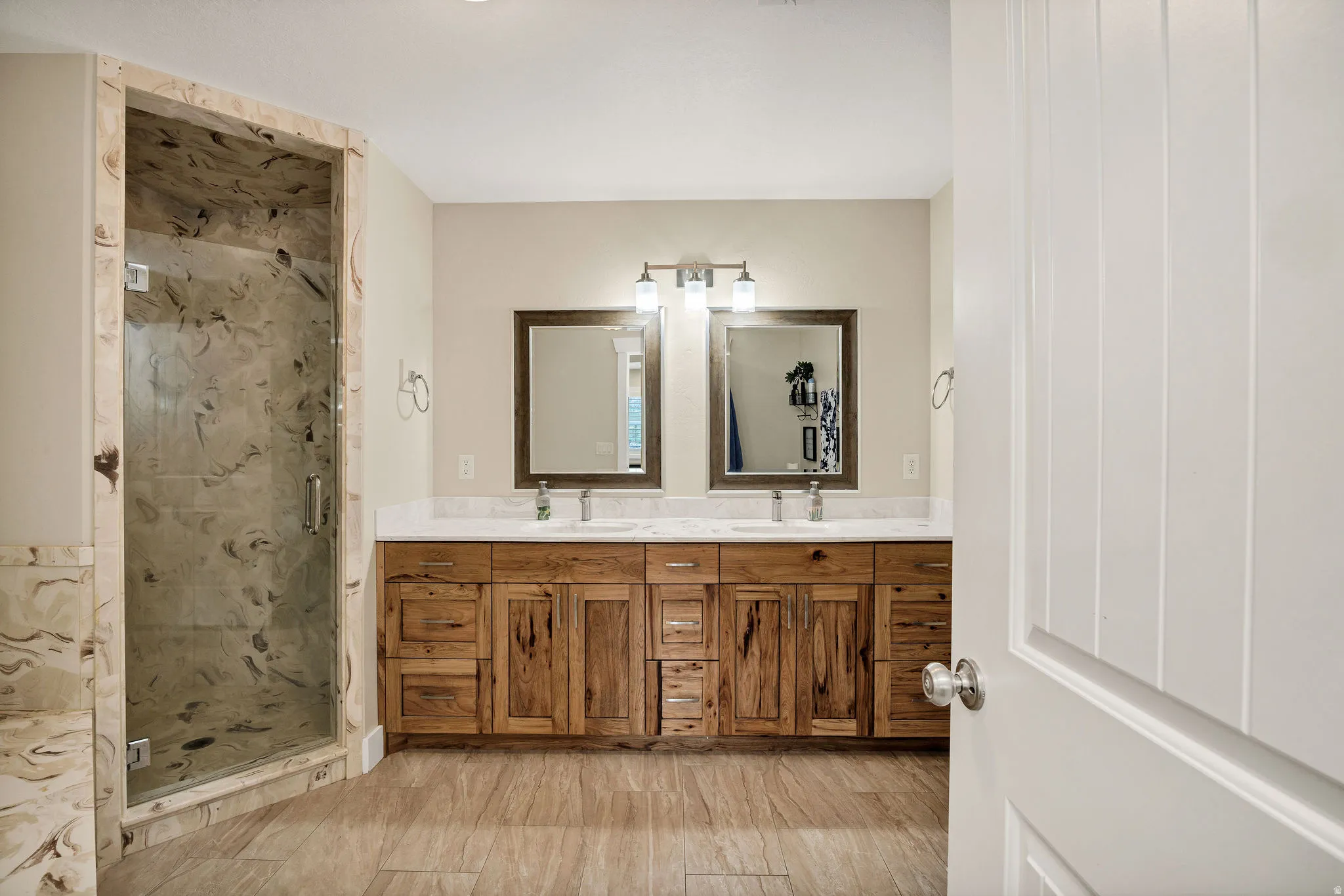Spacious primary bathroom featuring a glass-enclosed shower with stone-look tile surround, a double vanity with distressed wood-finish cabinetry, a white countertop, and wood-finish tile flooring