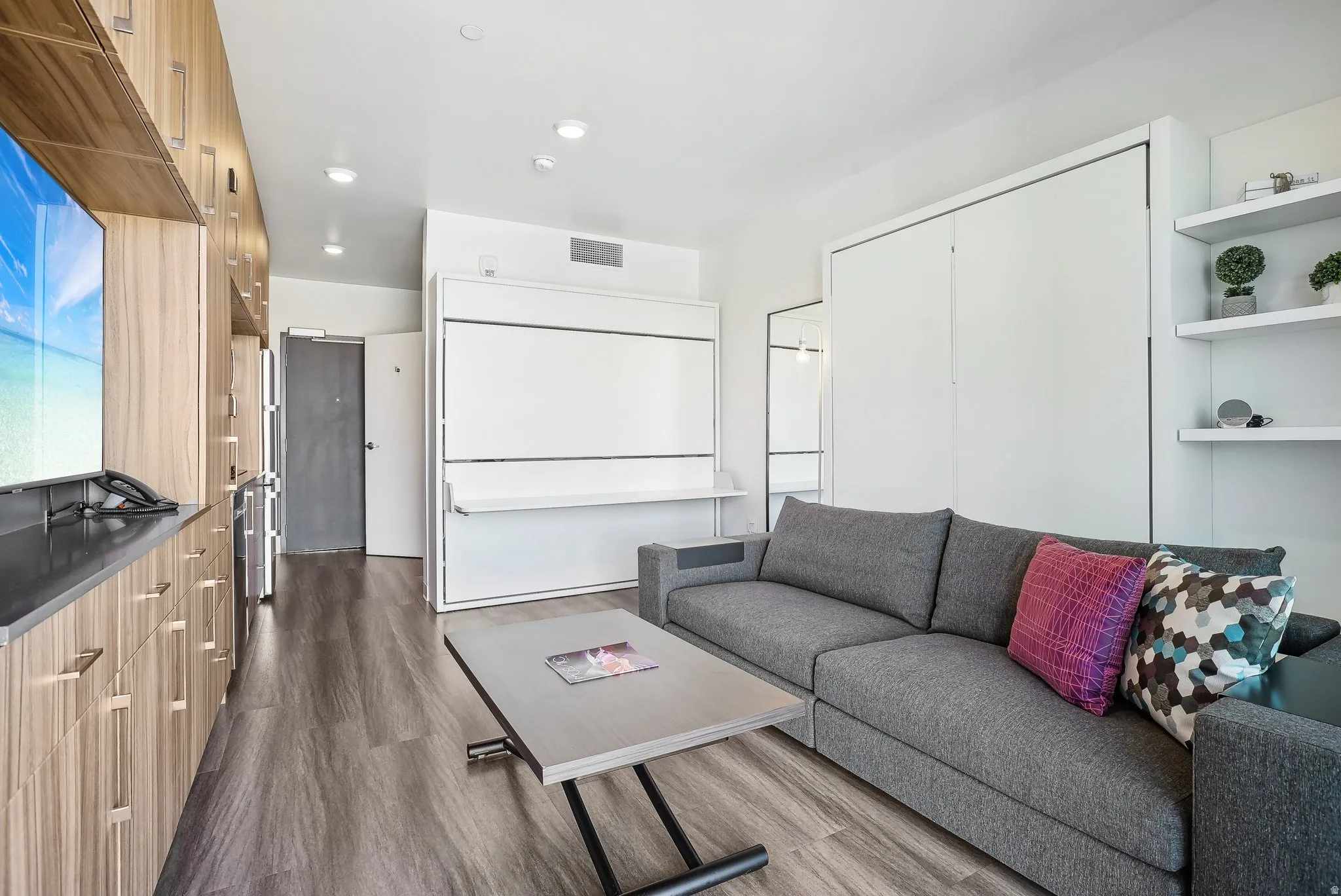 Open-concept living space featuring wood-finish flooring, a built-in wall unit with integrated cabinetry, a versatile murphy bed system, and floating wall shelves