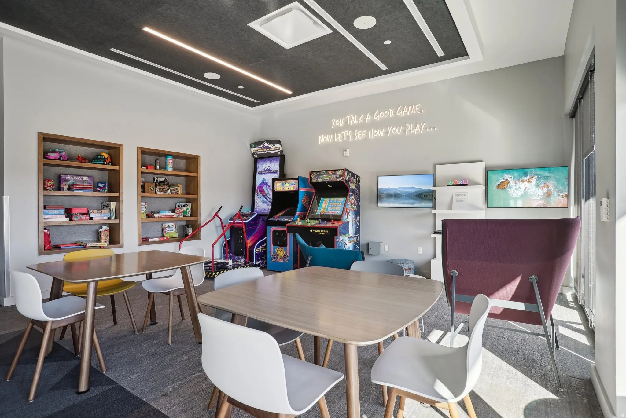 Recreational room featuring a dark ceiling with linear lighting, recessed wall shelving, two arcade machines, and dual wall-mounted displays