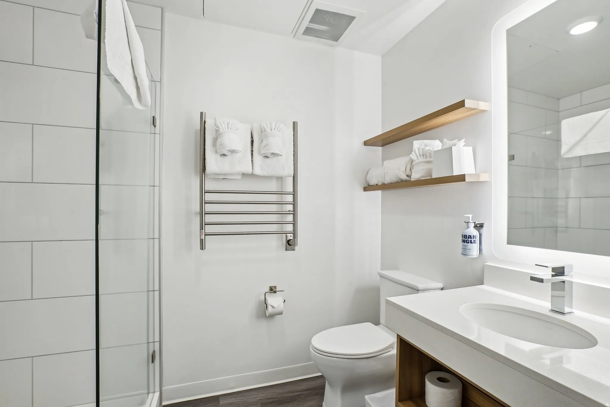 Bathroom featuring a glass-enclosed shower with white subway tile, a vanity with an integrated sink and chrome faucet, an illuminated mirror, and floating wood shelves