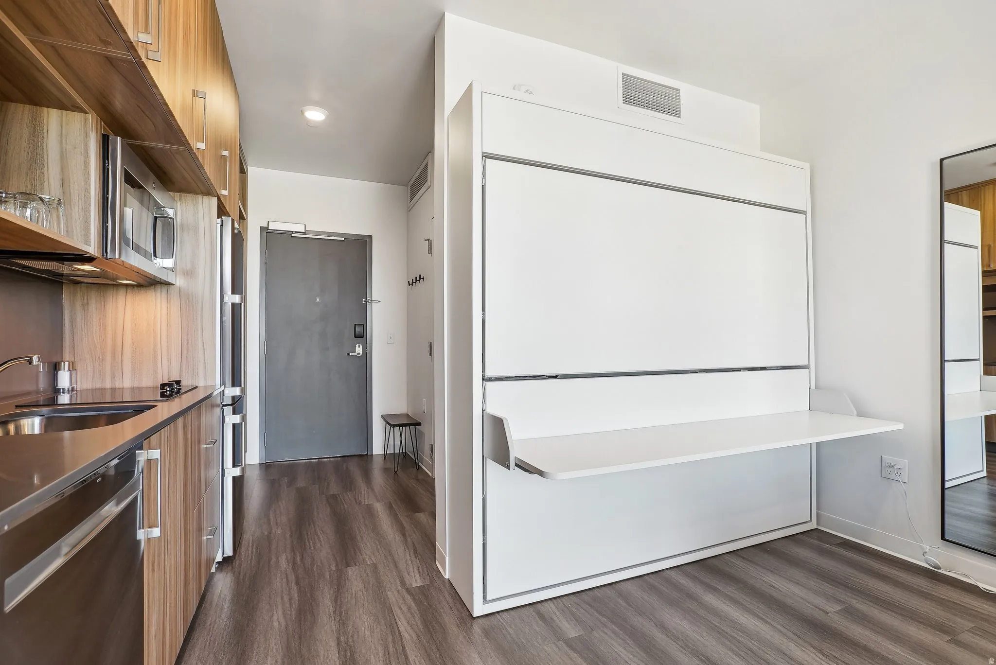 Compact living area featuring a built-in wall bed with an integrated desk, wood-finish flooring, and a stainless steel kitchen with a microwave and refrigerator